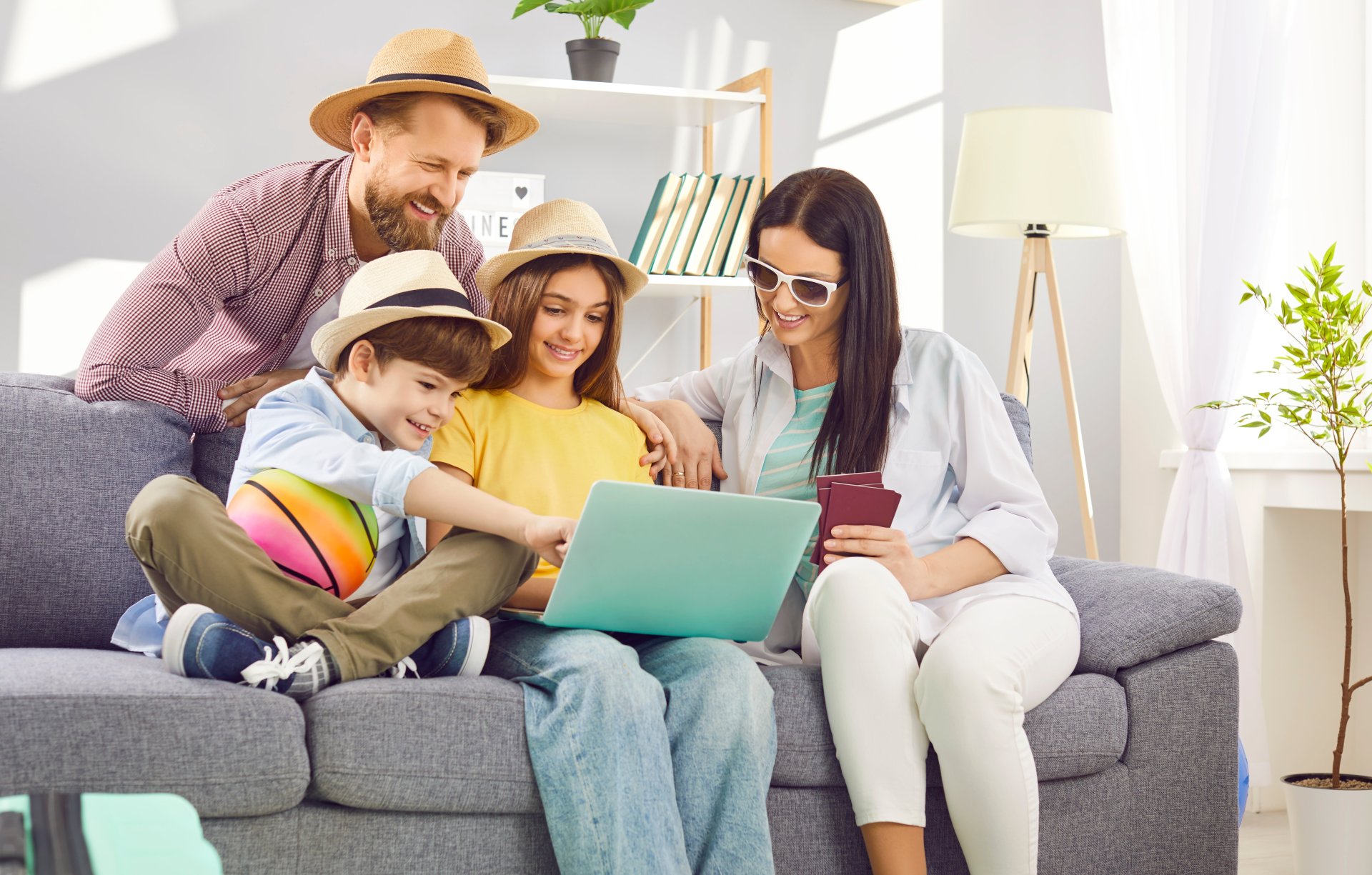 O familie asezata pe canapea in living foloseste un laptop pentru a planifica vacanta de vara, verificand oferte de zboruri si hoteluri in timp ce copiii stau langa ei si privesc curiosi, intr-o atmosfera relaxata si entuziasta de pregatire a calatoriei