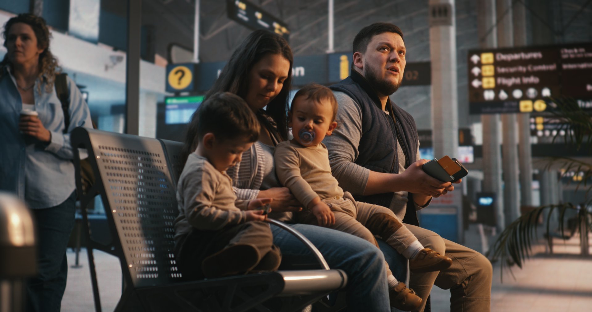 O familie de patru persoane asezata in terminalul unui aeroport, cu bagaje langa ei, in timp ce tatal verifica pe smartphone biletele de imbarcare digitale, iar mama are grija de doi copii mici inainte de zbor.