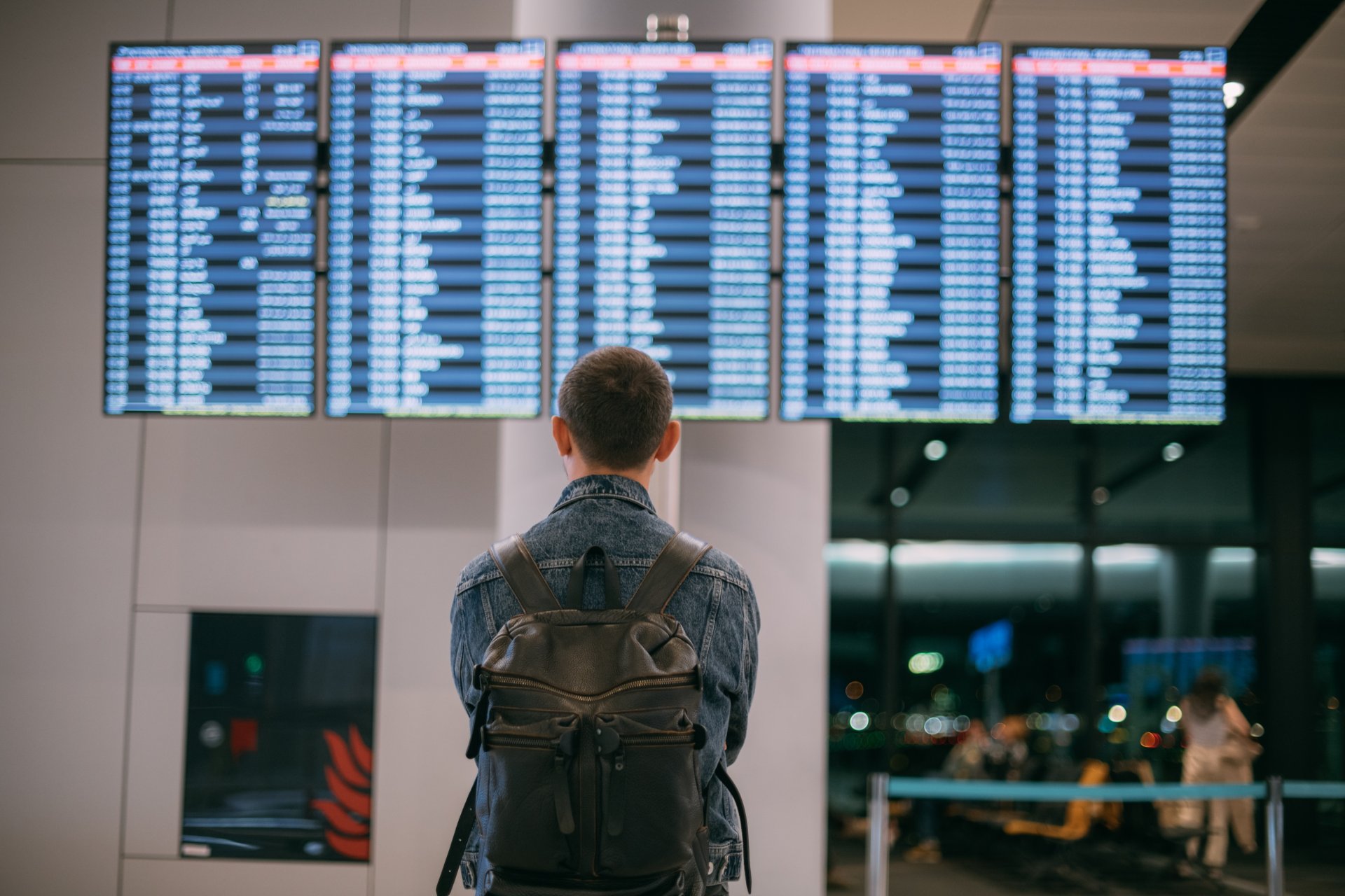 Un tanar cu rucsac si bagaj de mana sta in terminalul unui aeroport modern si priveste un ecran mare cu plecari si sosiri. Atmosfera este de asteptare si miscare, cu luminile reci ale terminalului si alti pasageri in fundal care se deplaseaza rapid.