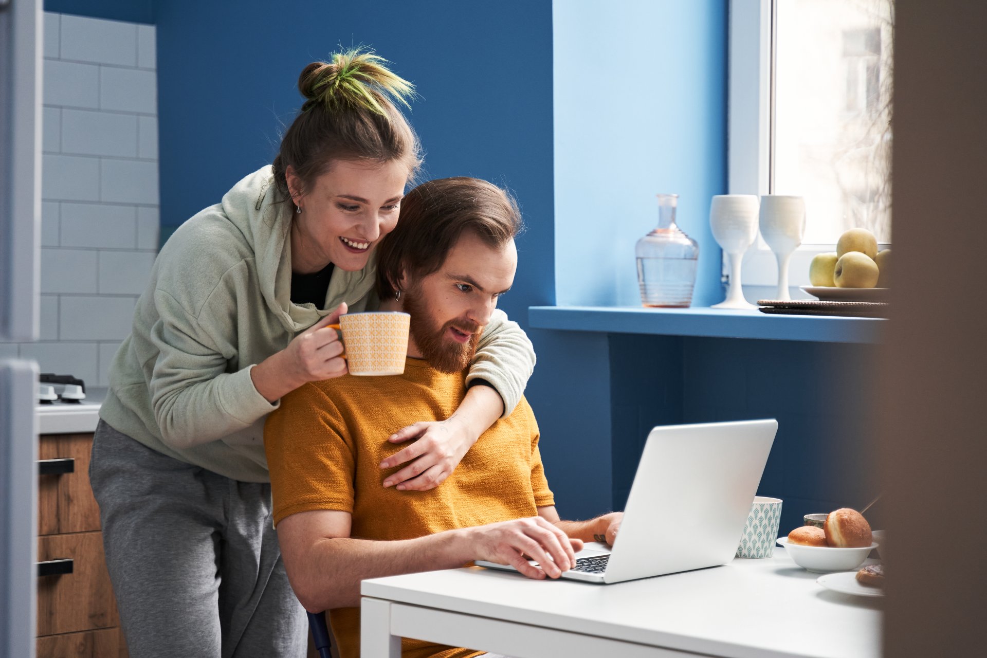 Un cuplu relaxat sta pe canapea acasa, femeia foloseste un laptop in timp ce bea ceai, iar partenerul ei cu barba o imbratiseaza din lateral. Atmosfera este calda si intima, cu un sentiment de confort si liniste intr-un moment de pauza impreuna.