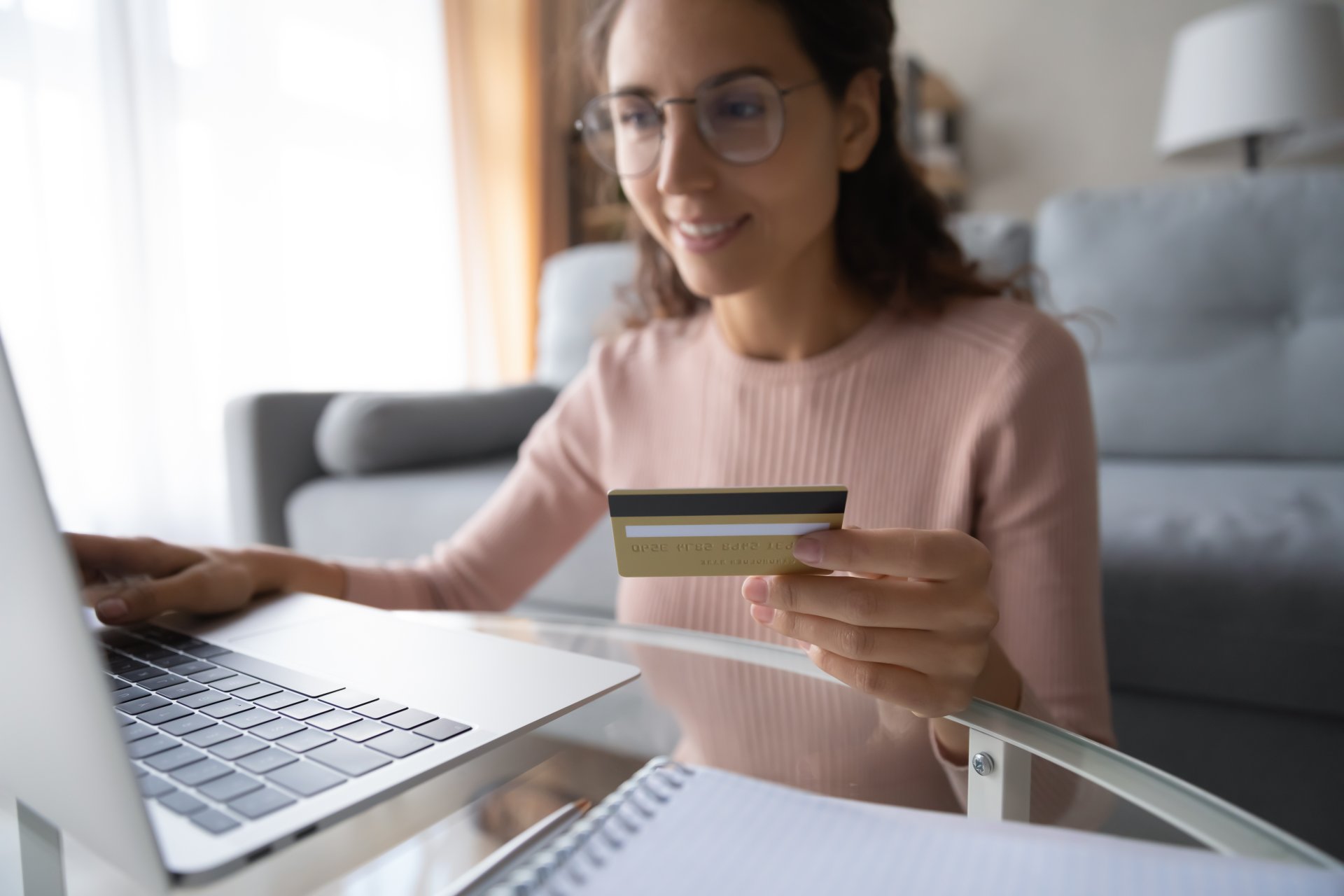 O femeie zambitoare cu ochelari tine un card bancar in mana in timp ce introduce datele de plata pe un laptop, intr-un mediu relaxat de acasa, sugerand o sesiune de cumparaturi online sigura si confortabila