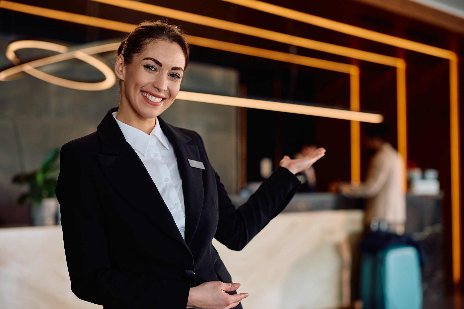 Un receptioner de hotel zambitor sta la receptie si intampina oaspetii, intr-un lobby modern si luminos, cu atmosfera profesionala si primitoare, sugerand servicii de ospitalitate de calitate intr-un hotel international