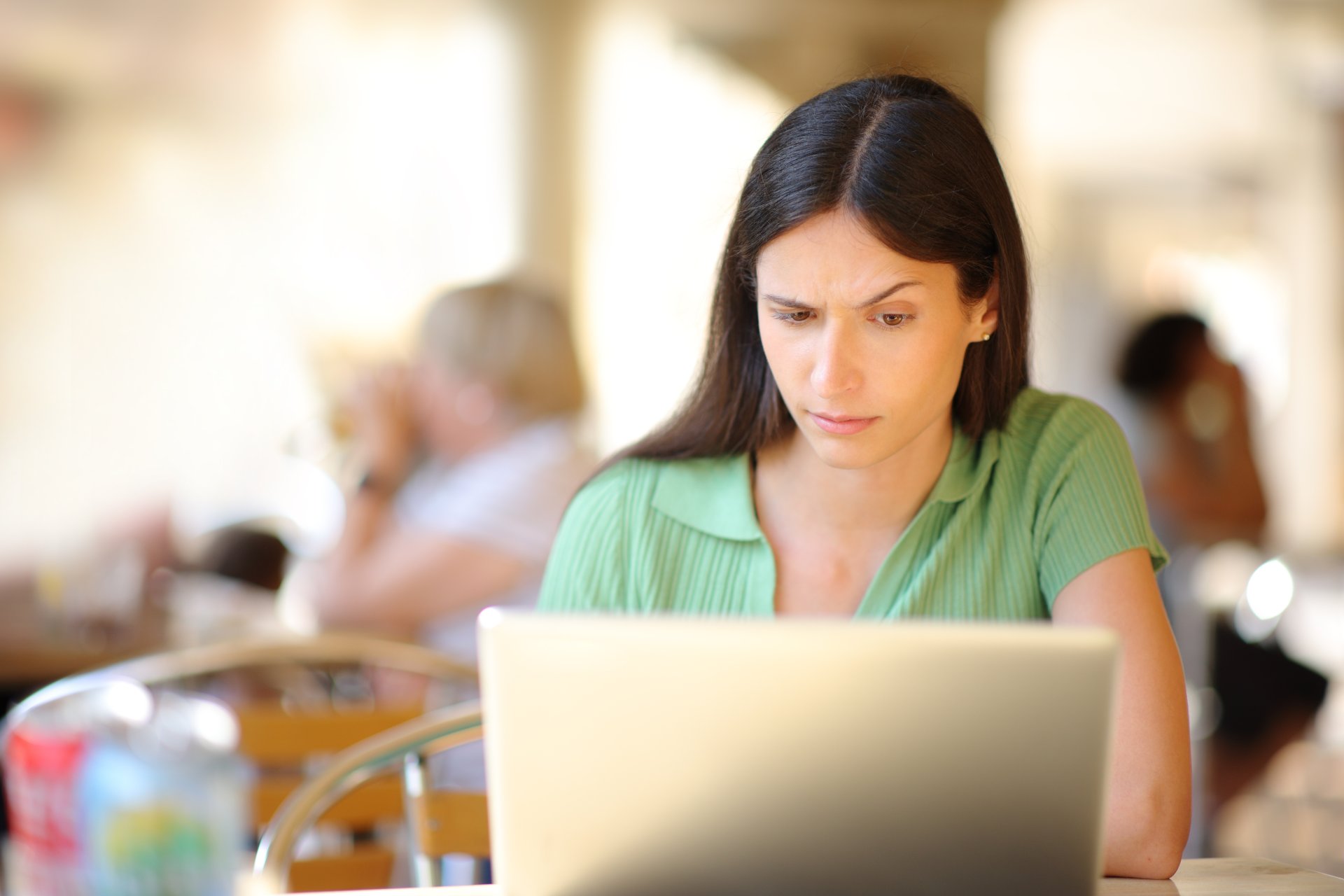 O femeie sta la o masa pe terasa unui restaurant si foloseste un laptop, avand o expresie usor suspicioasa si concentrata. In jur se vad mese de cafenea si o atmosfera urbana relaxata, cu lumina naturala de zi.