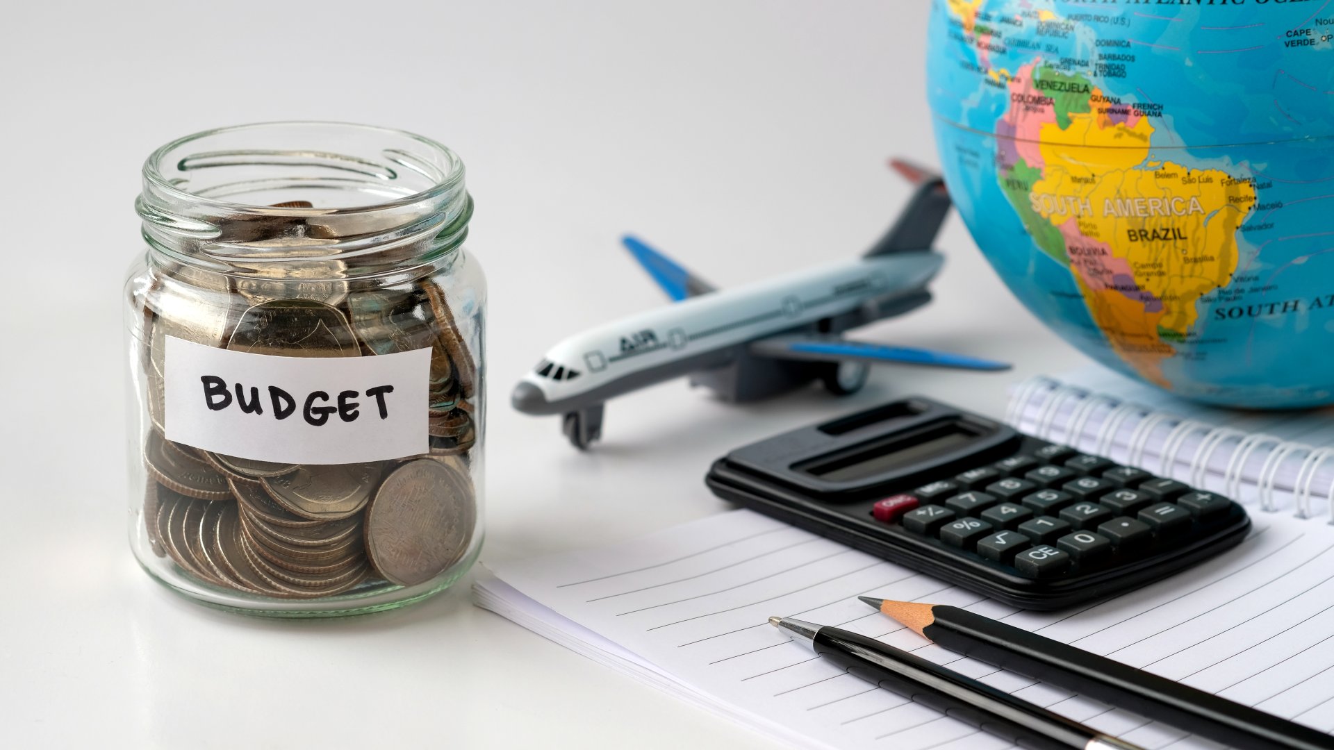 Masa de birou cu un borcan plin de monede, un calculator si un glob pamantesc, alaturi de un avion de jucarie care sugereaza planificarea unei vacante si economisirea banilor pentru calatorii internationale