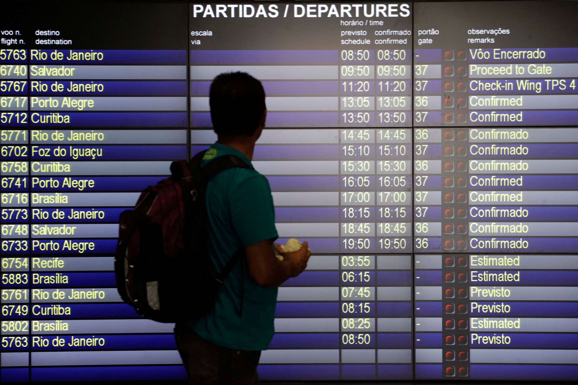 Un calator sta in fata unui panou mare de plecari din aeroport si cauta informatii despre zboruri. In jurul lui se vede un terminal modern, cu oameni care se grabesc si lumini specifice unui aeroport aglomerat.