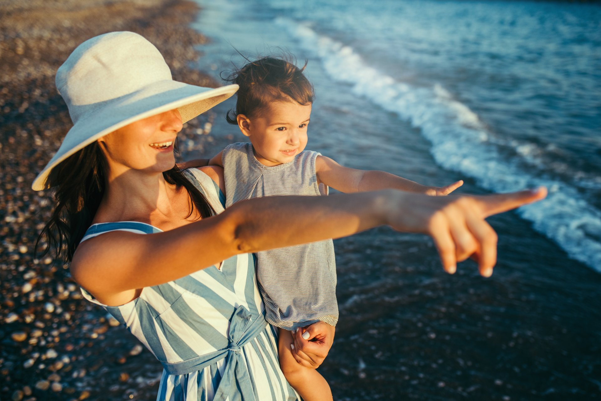 O mama si fiica ei se imbratiseaza pe malul marii, pe o plaja insorita, in timpul unei vacante de vara. Mama poarta palarie si arata ceva in departare, in timp ce copilul priveste spre orizont, intr-un cadru relaxat si fericit de familie in natura