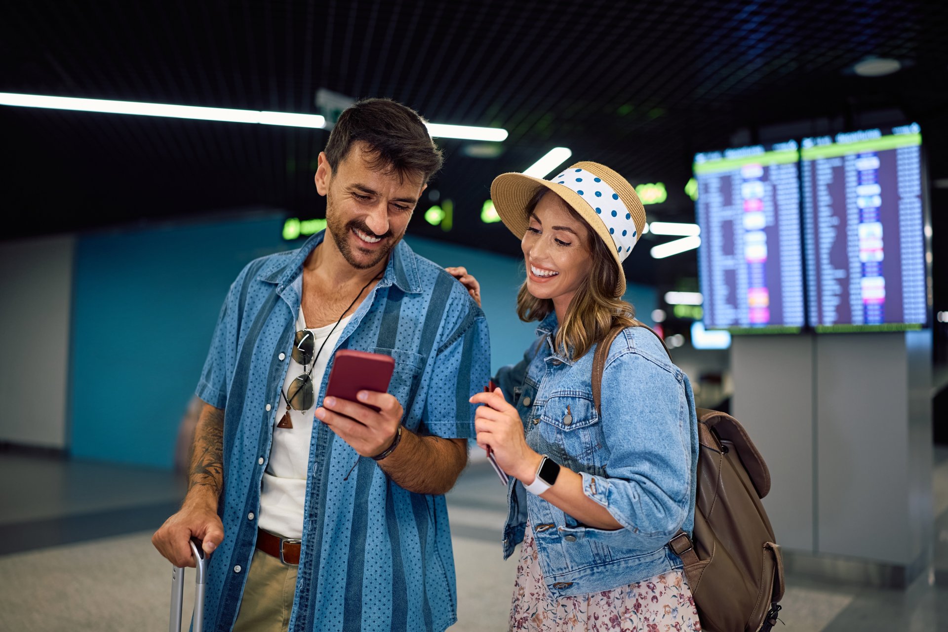 Un cuplu zambeste si foloseste telefonul mobil in zona de plecari a aeroportului, in timp ce asteapta imbarcarea pentru urmatorul zbor.