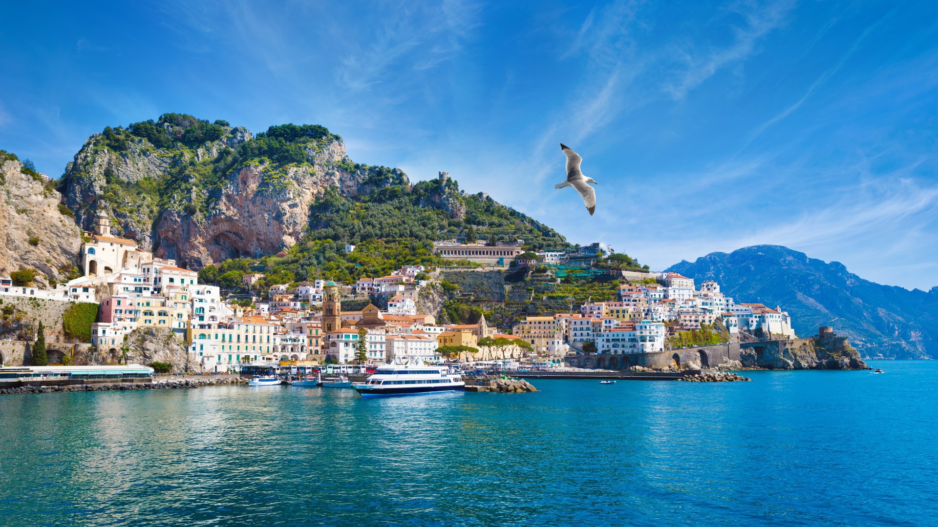 Panorama asupra orasului Amalfi, cu case colorate construite pe versant, portul la malul Marii Mediterane si stancile spectaculoase ale Coastei Amalfitane sub un cer senin.