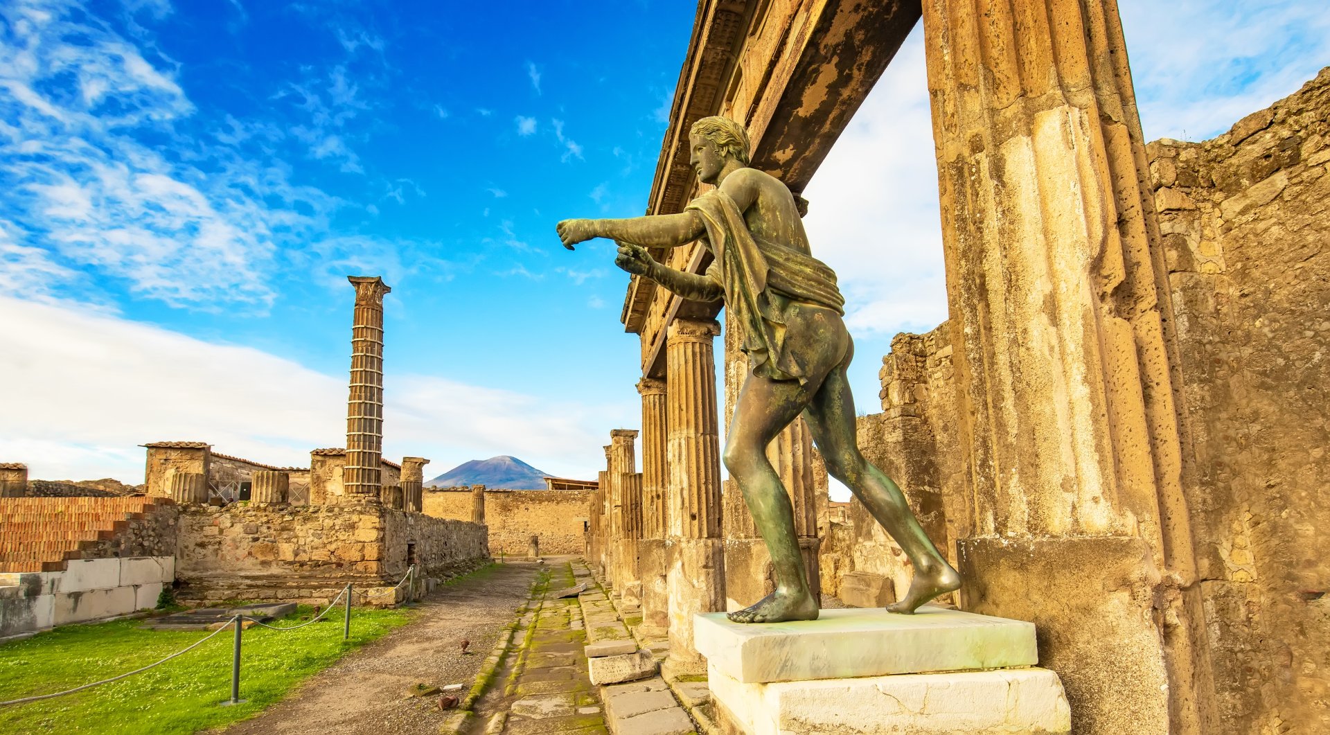 Statuia din bronz a lui Apollo domina ruinele orasului antic Pompei, cu vestigiile istorice si muntii din fundal, intr-unul dintre cele mai cunoscute situri arheologice din Italia.