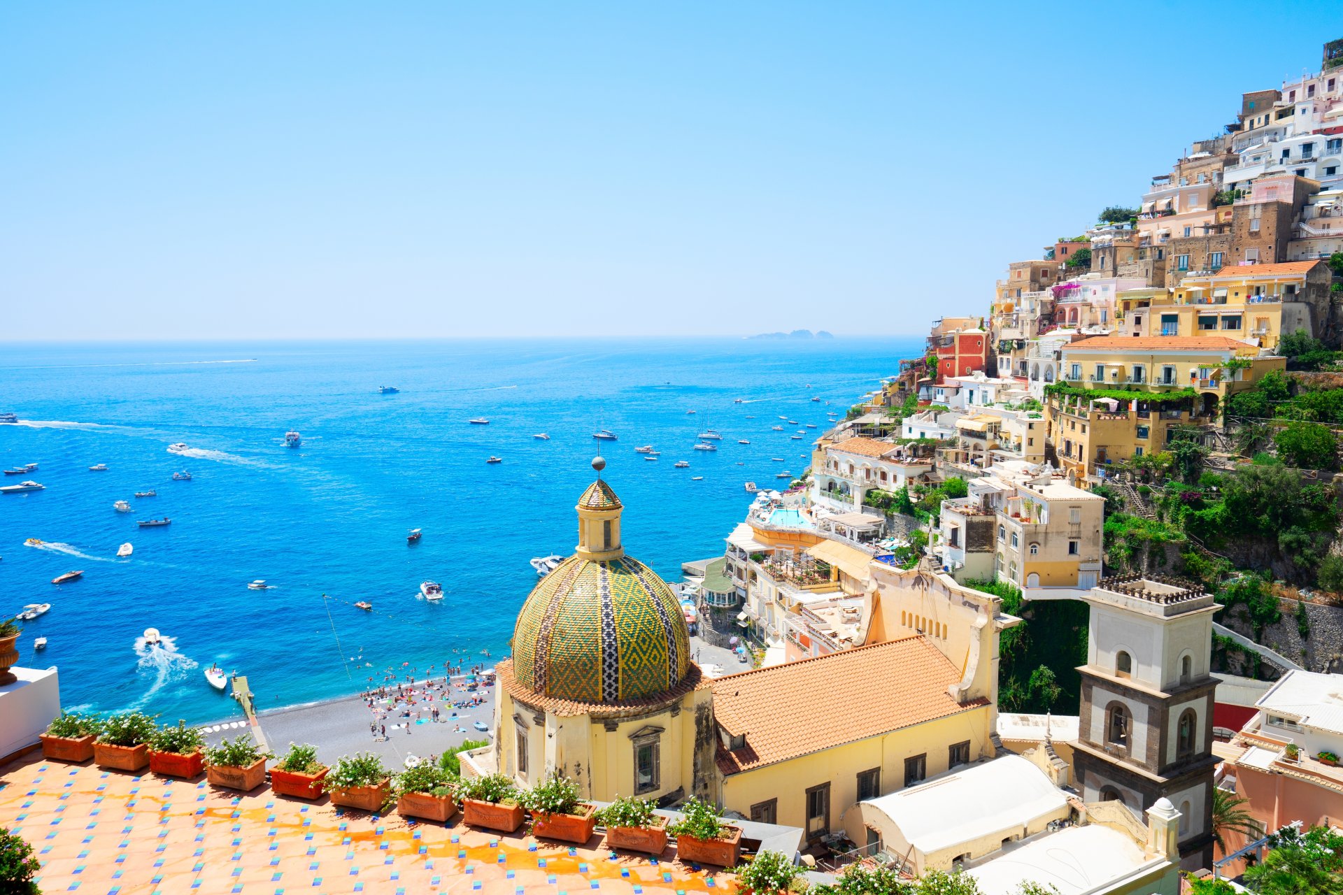 Panorama asupra statiunii Positano, cu case colorate construite pe versant, plaja la malul Marii Tireniene si ambarcatiuni ancorate in larg, intr-o zi senina de vara pe Coasta Amalfitana.