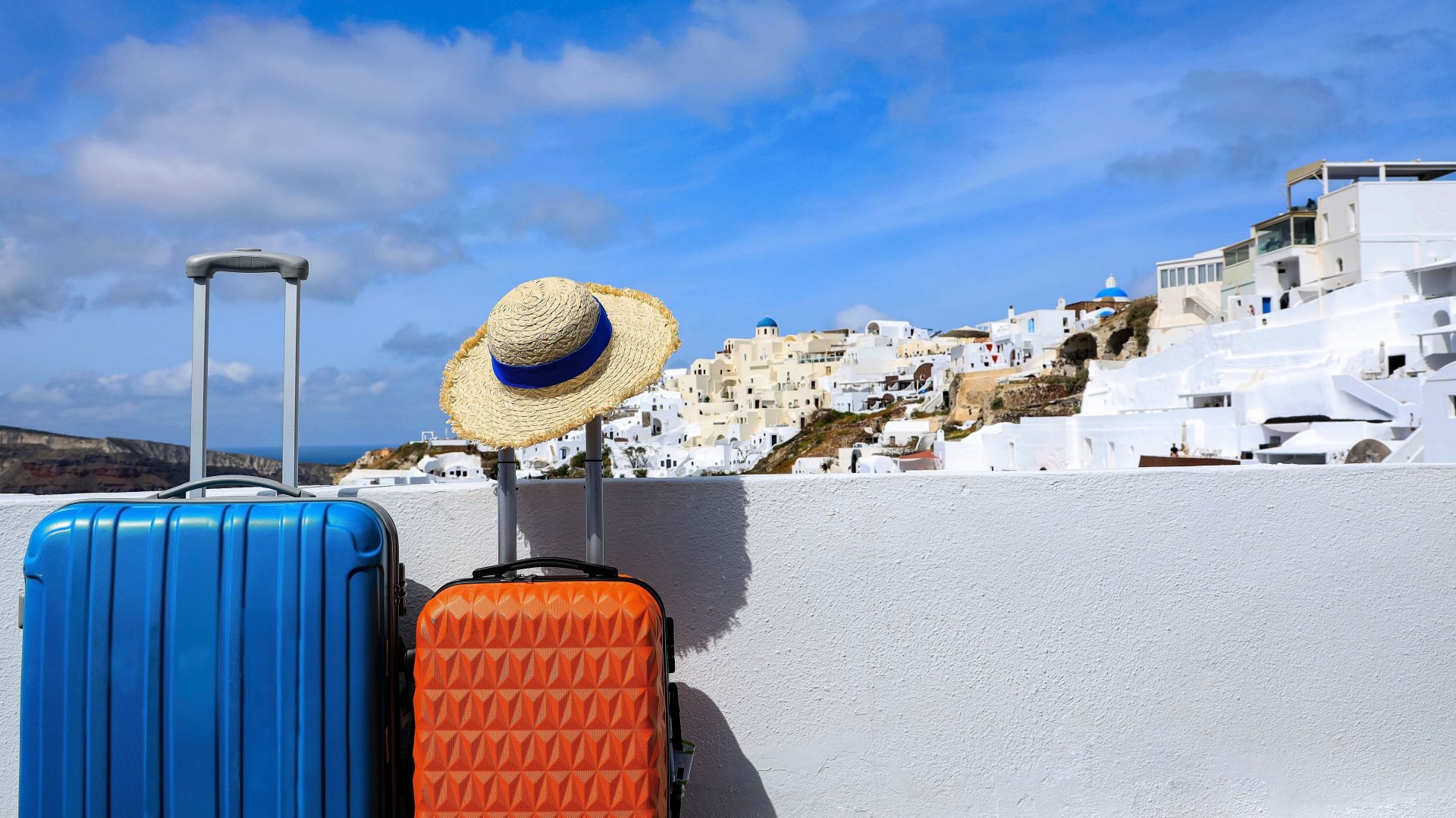 Doua valize si o palarie sunt asezate in prim-plan, avand in fundal casele albe si cladirile specifice din Oia, Santorini, sub un cer albastru senin de vara.