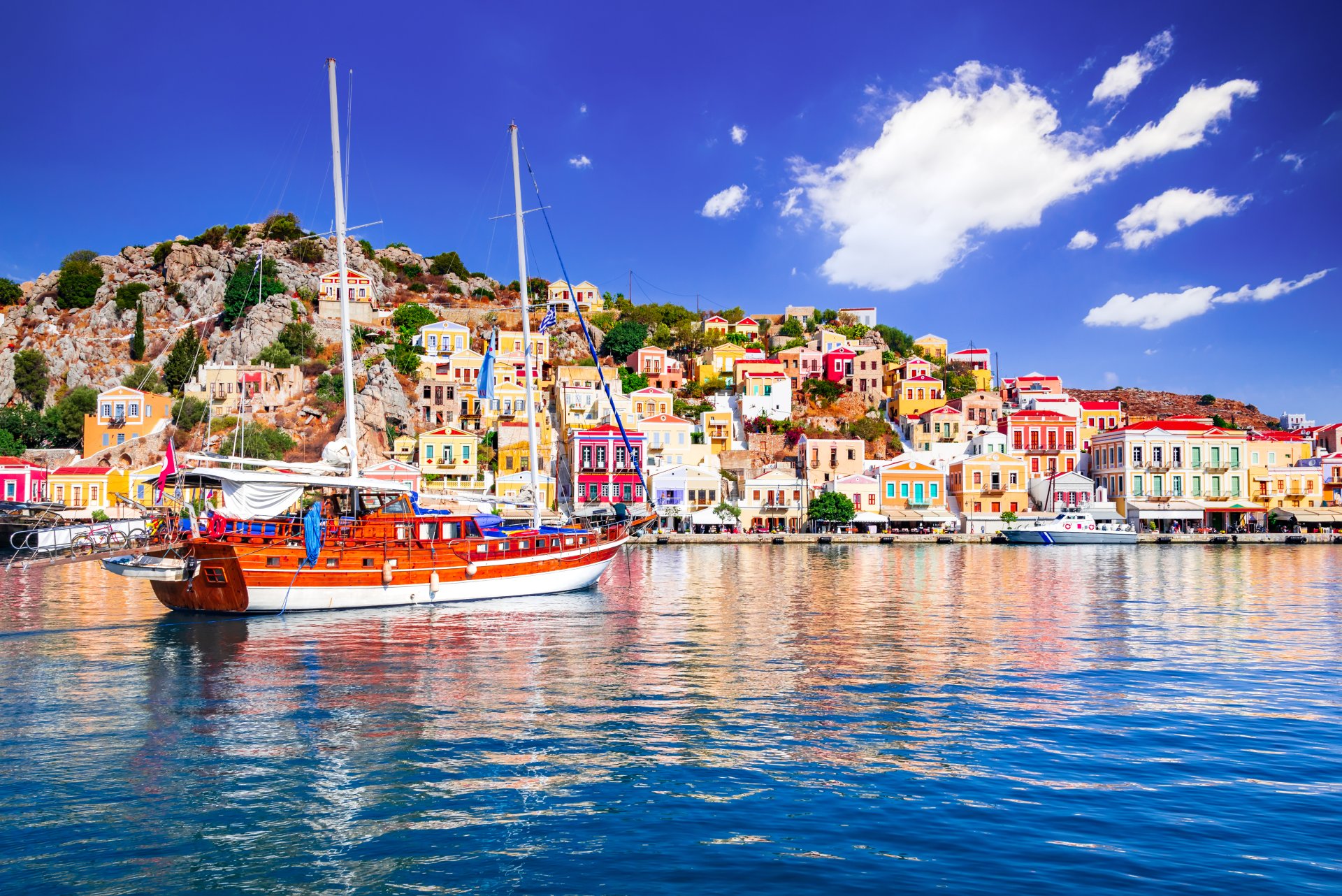 Vedere asupra golfului din insula Symi, cu case neoclasice colorate construite in amfiteatru pe dealuri, barci ancorate in port si ape linistite ale Marii Egee intr-o zi insorita de vacanta.