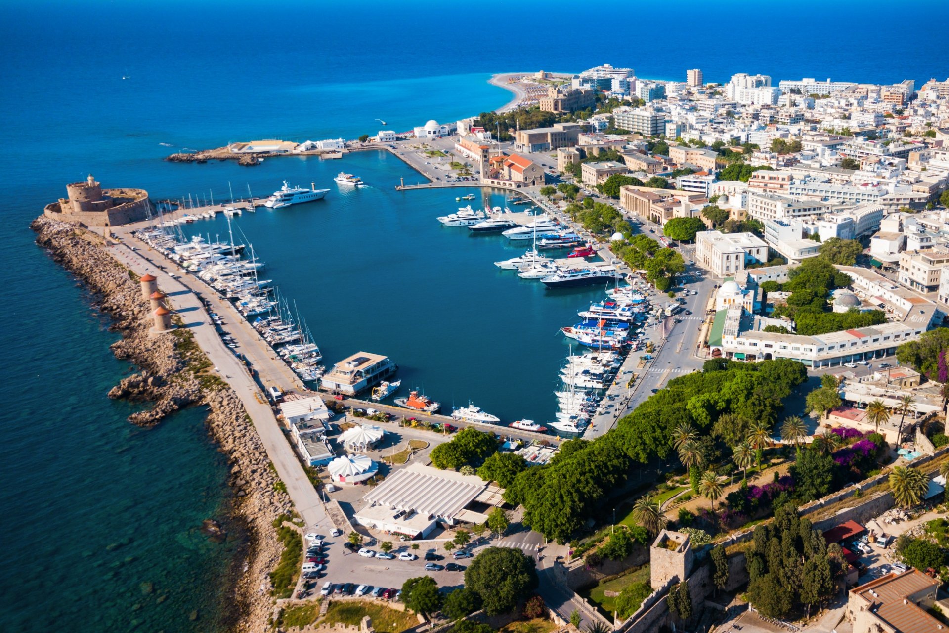 Vedere aeriana panoramica asupra portului Mandraki din orasul Rhodos, cu ambarcatiuni ancorate, cladiri de-a lungul falezei si Marea Egee in fundal.