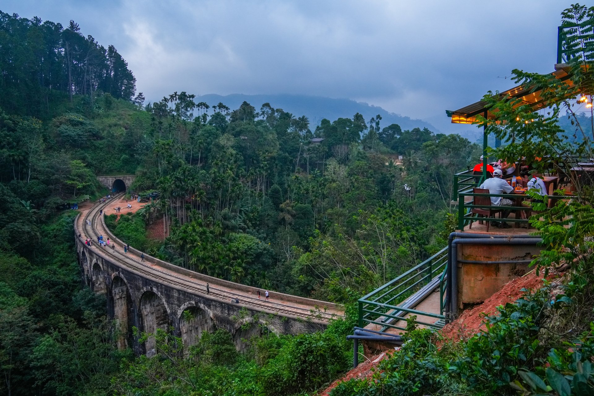 Pod de piatra cu noua arcade in zona muntoasa din Ella, Sri Lanka, inconjurat de vegetatie tropicala deasa si dealuri verzi, cu un tren care traverseaza lent podul intr-un peisaj pitoresc