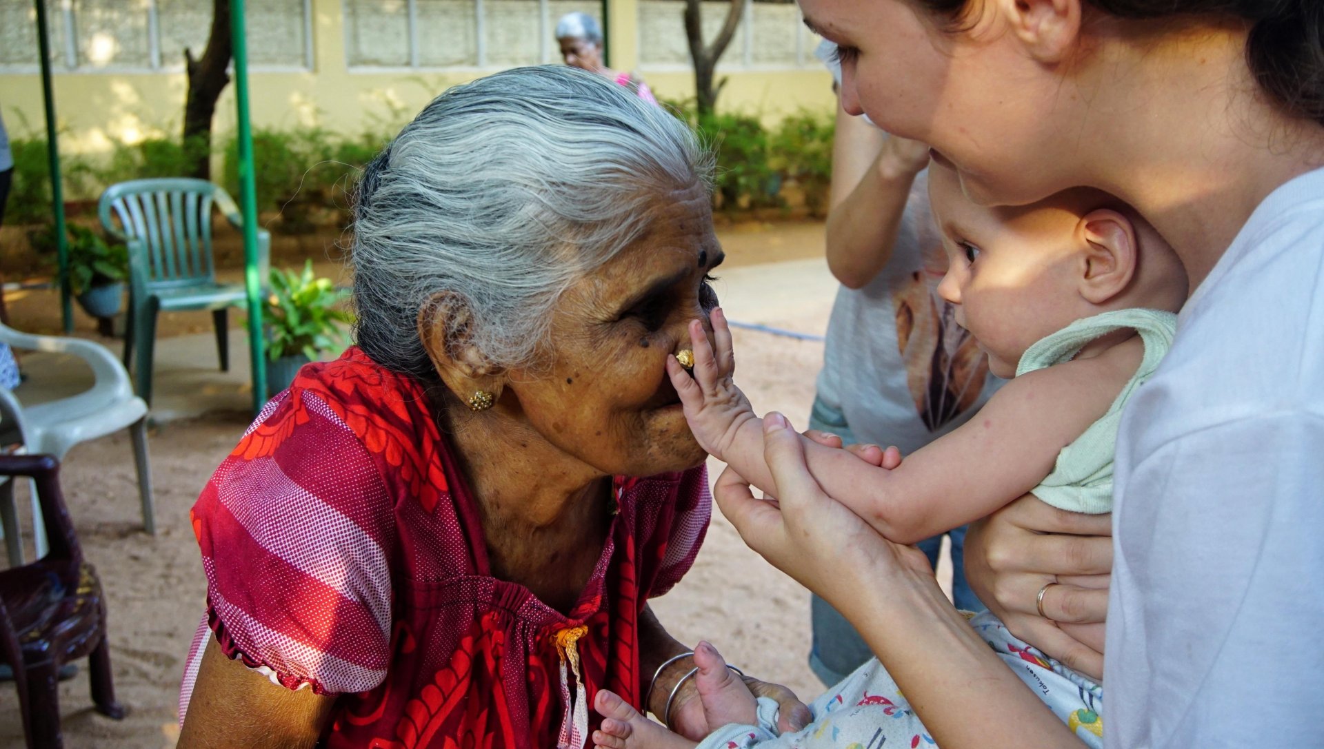 O femeie in varsta imbracata in rochie rosie traditionala sta intr-un azil de batrani din Uppuveli, Sri Lanka, in timp ce un copil mic se intinde spre ea pentru a-i atinge fata, intr-un moment emotionant de conexiune intre generatii, surprins in aer liber intr-un cadru simplu si cald