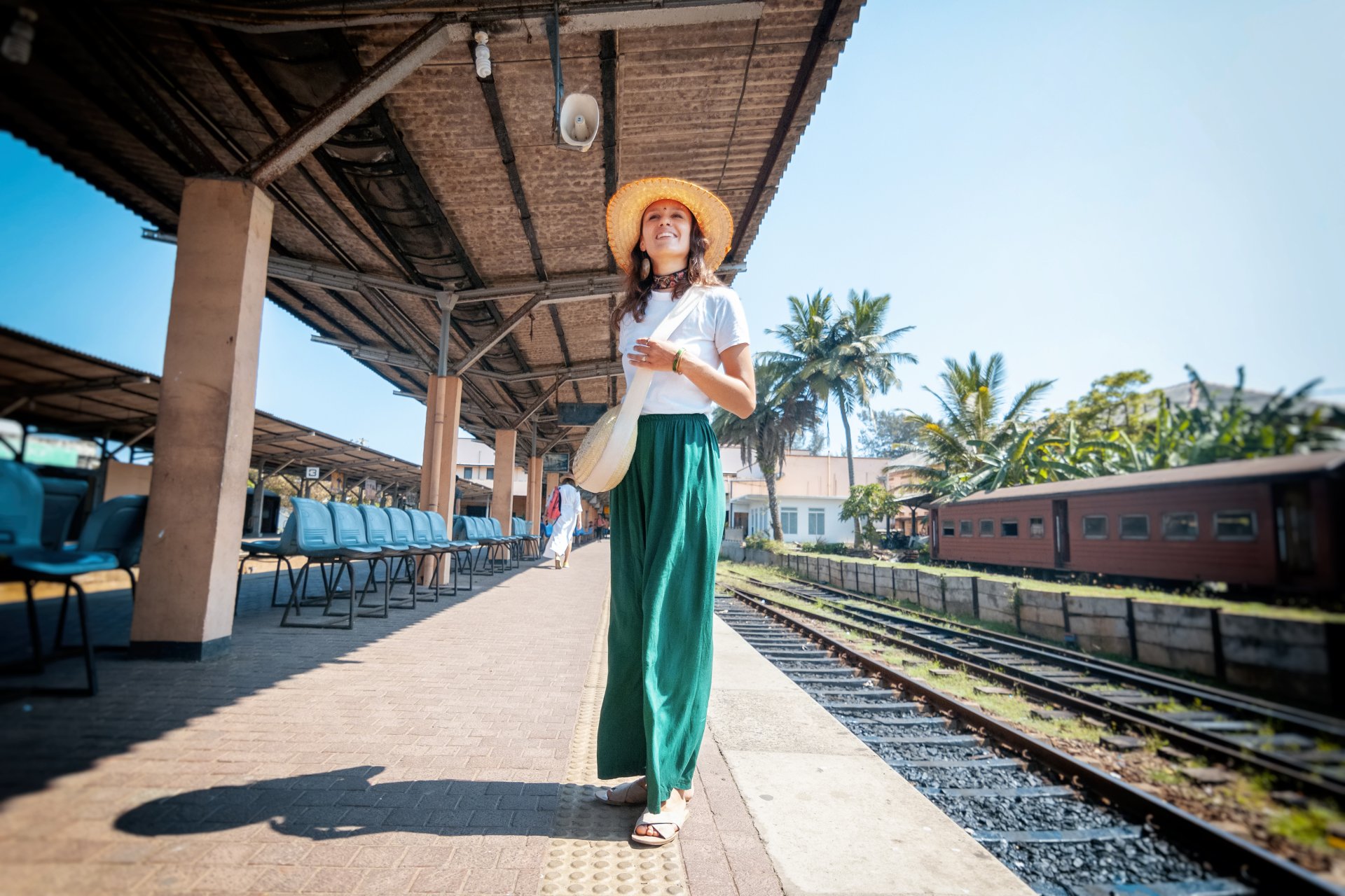 O tanara calatoare cu palarie de vara sta pe un peron de gara in Sri Lanka, asteptand un tren, cu o atmosfera tropicala si exotica in jur