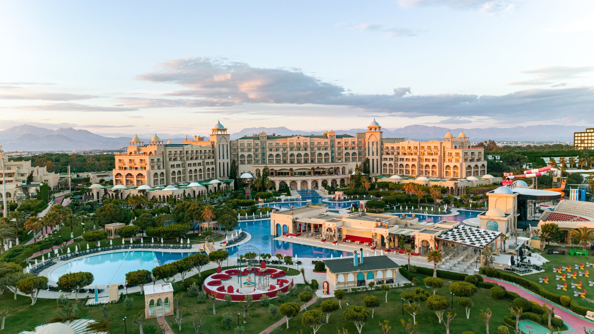 Vedere cu un hotel mare de lux pe malul marii in Antalya, cu piscina exterioara, sezlonguri aliniate si turisti care se relaxeaza sub soare intr-o atmosfera de vacanta all inclusive