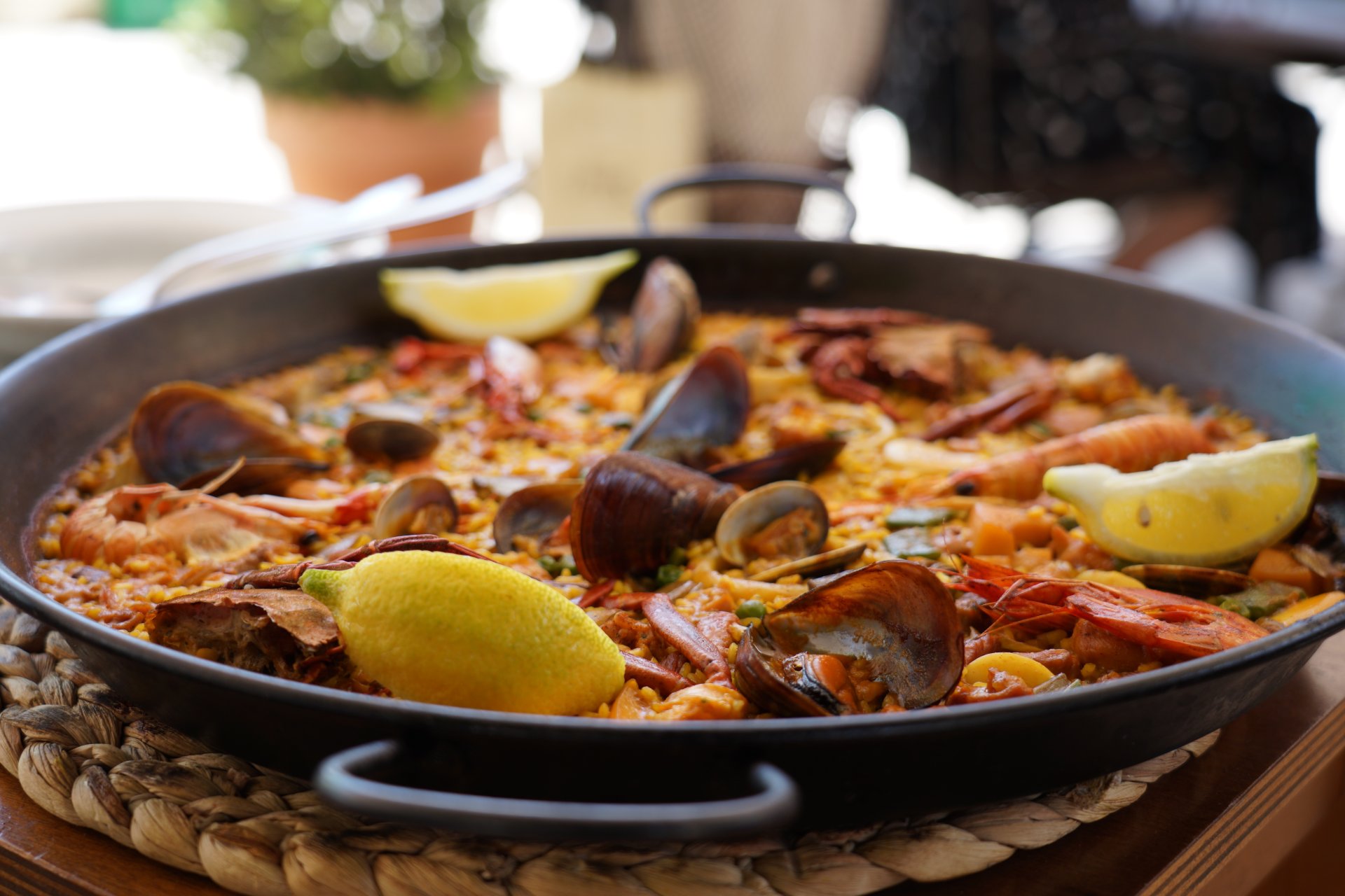 O paella servita intr-un restaurant de pe insula Mallorca, cu o tigaie mare plina de orez galben, fructe de mare si felii de lamaie, asezata pe o masa in aer liber unde se vede o atmosfera relaxata de vacanta cu turisti care mananca la terase langa mare