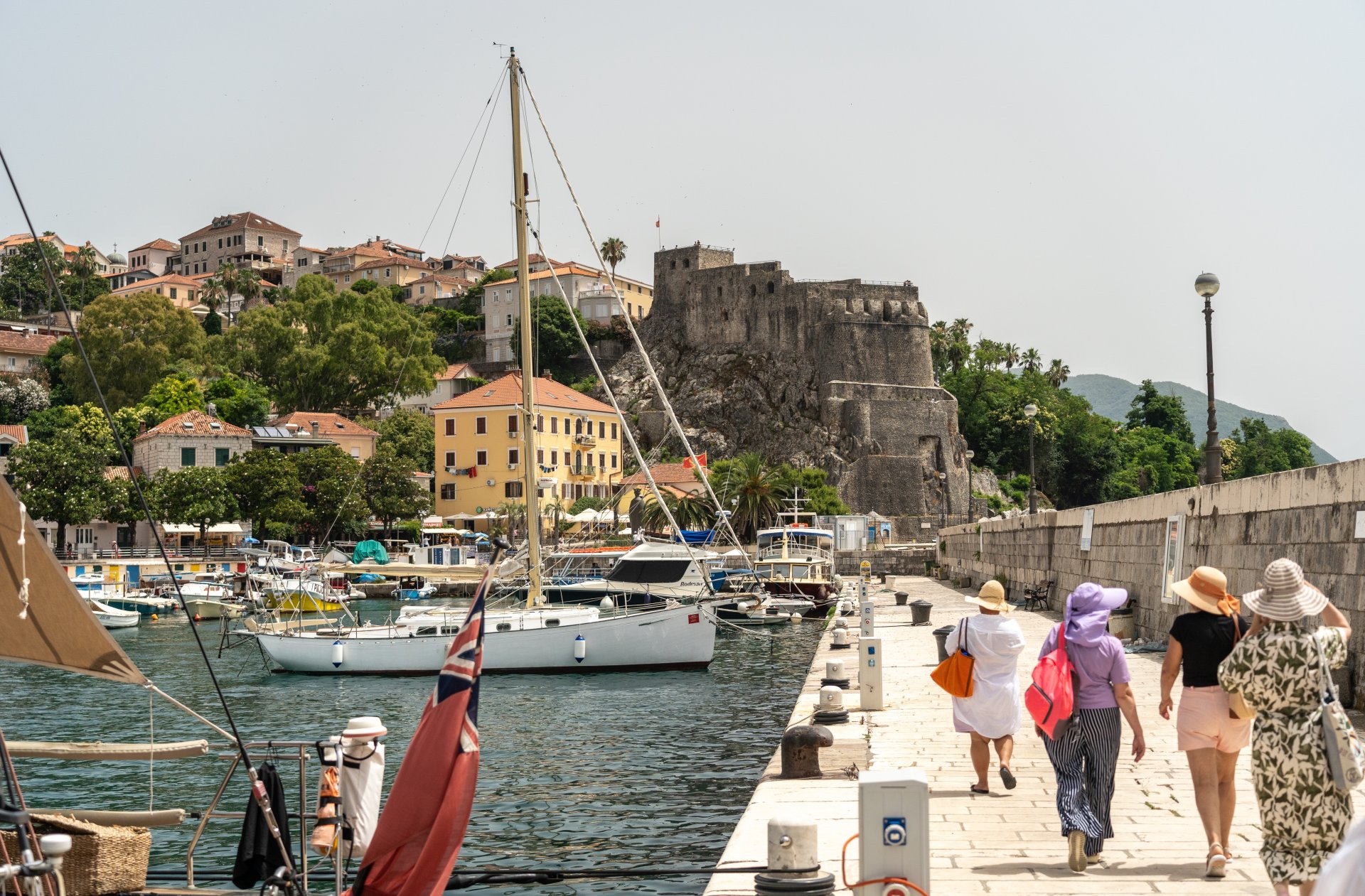 Barci turistice si iahturi ancorate in marina din Herceg Novi, pe coasta Adriaticii, intr-o zi de vara, cu promenade si cladiri specifice litoralului din Muntenegru pe fundal.