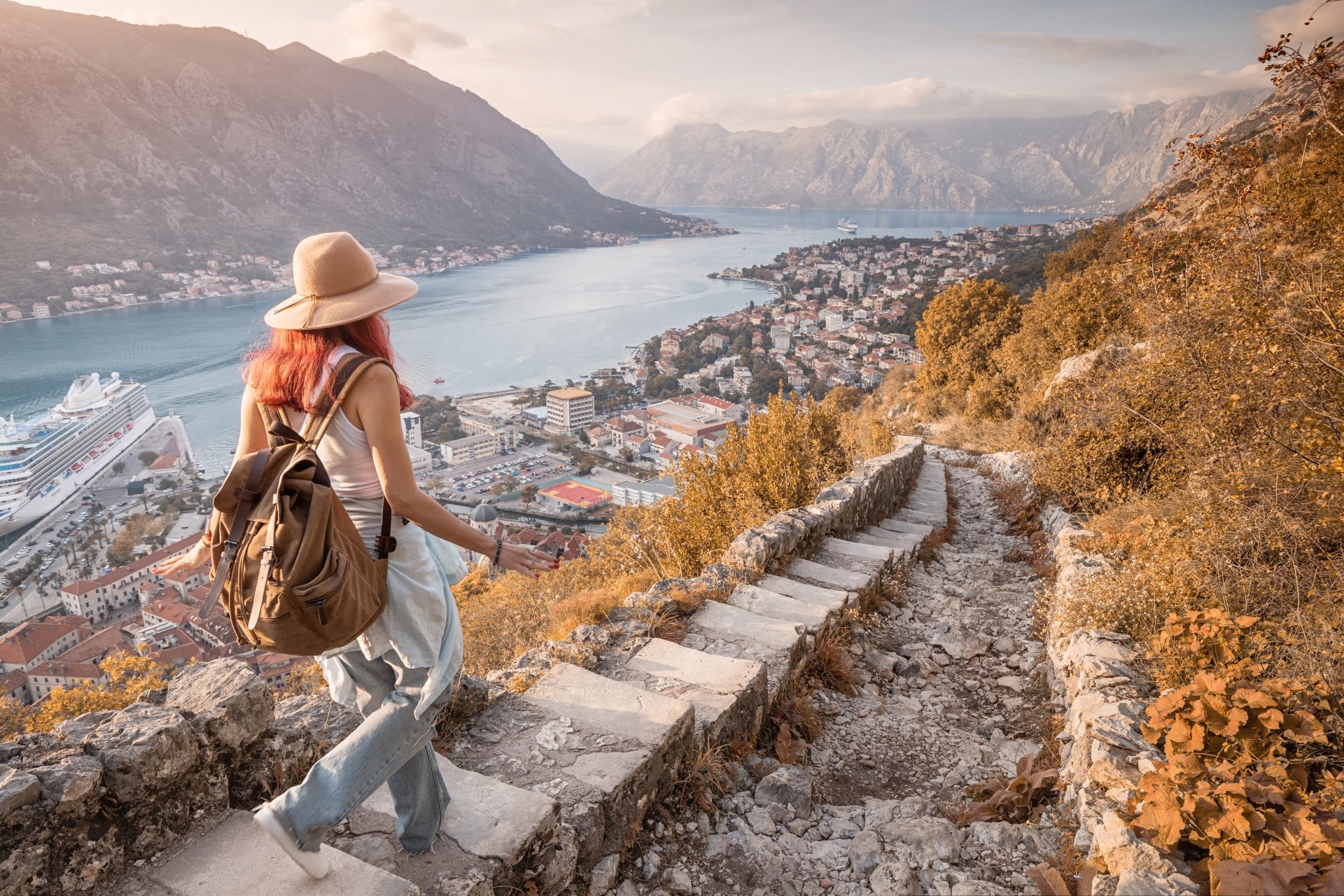 Turista coboara pe trepte din piatra si admira panorama spectaculoasa asupra Golfului Kotor la apus, inconjurat de munti si cladiri istorice din Muntenegru.