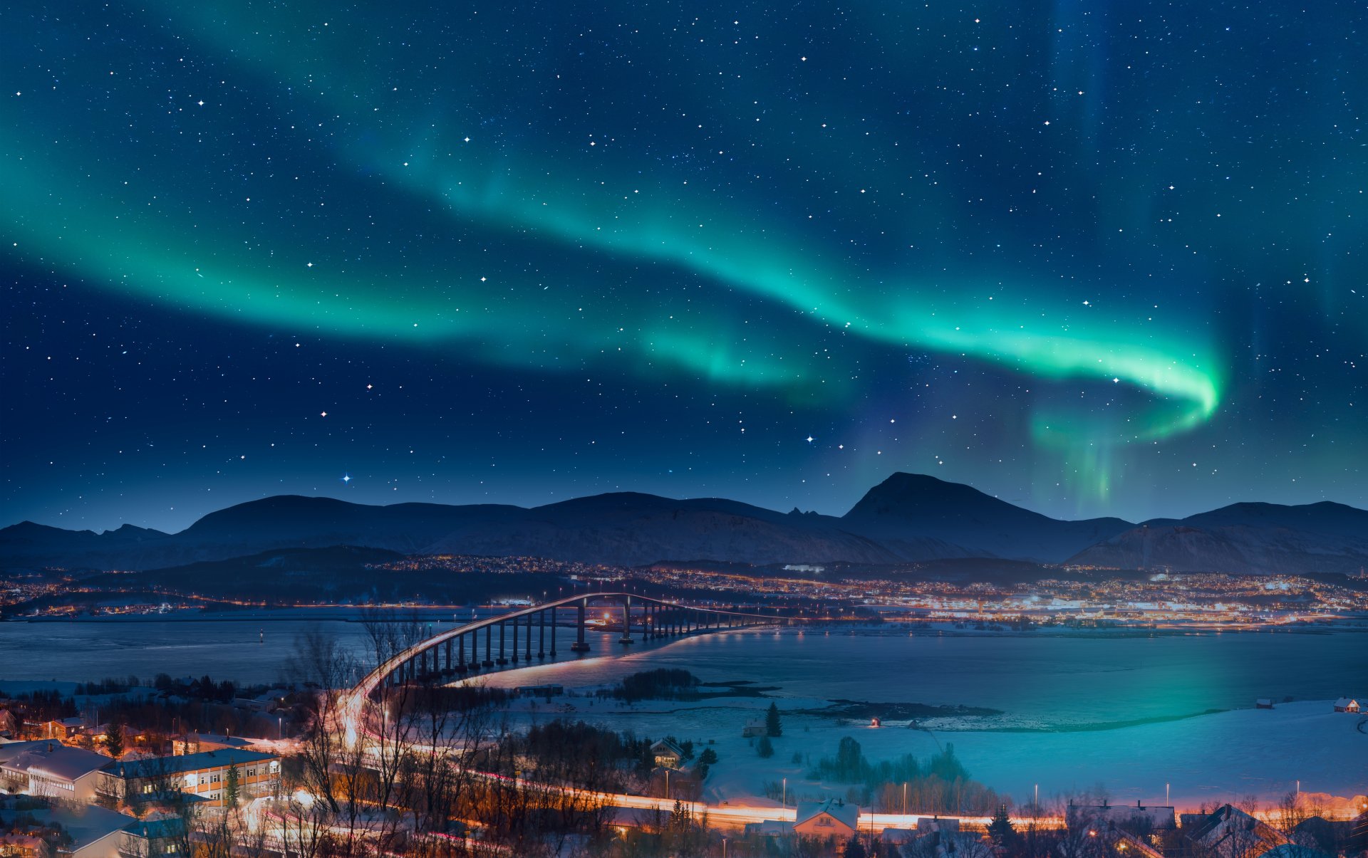 Aurora boreala lumineaza cerul deasupra orasului Tromso, iar Podul Sandnessundet se reflecta in apa, oferind un peisaj spectaculos din nordul Norvegiei.