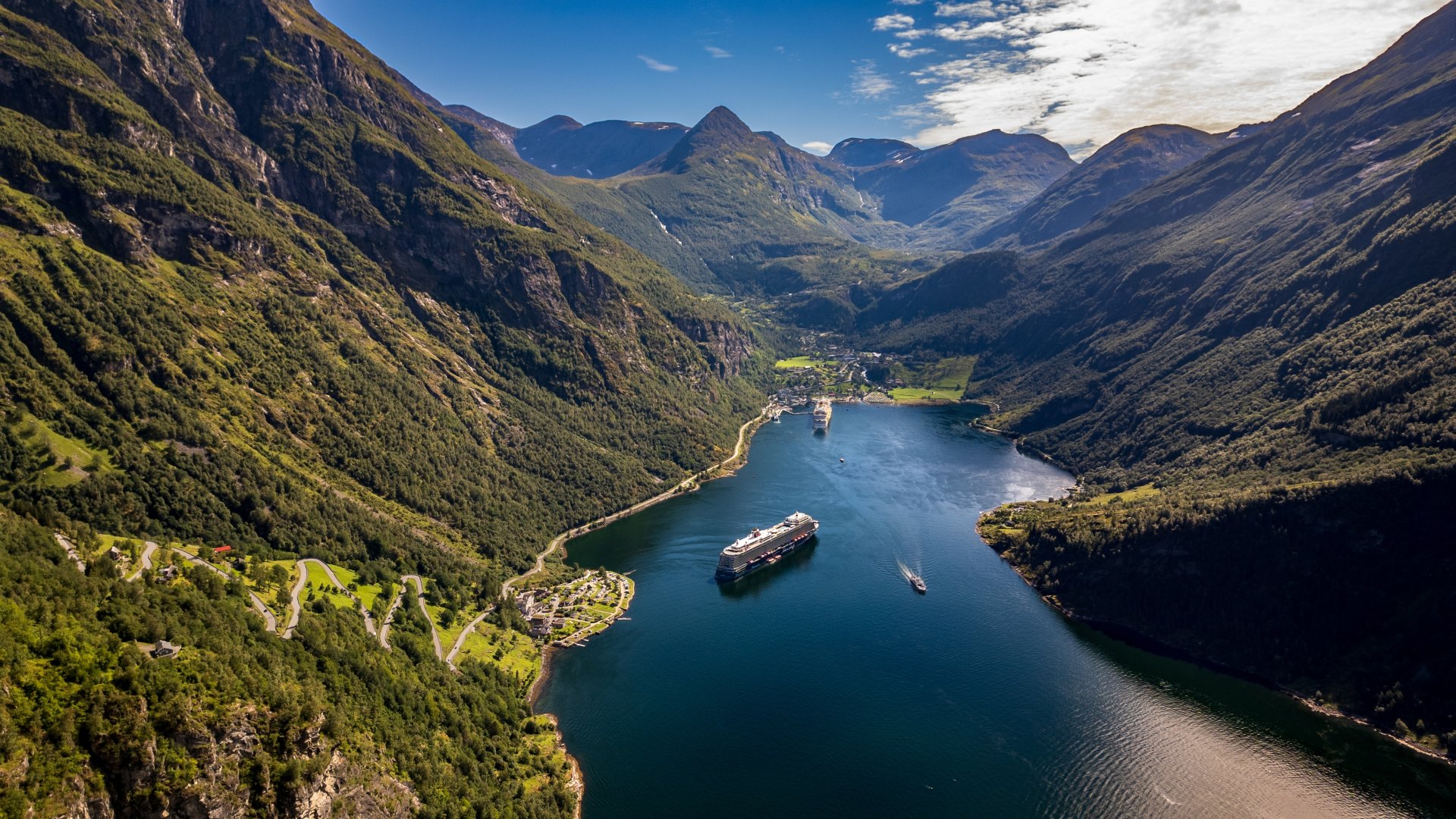 Vedere aeriana asupra fiordului Geiranger din Norvegia, cu nave de croaziera pe apa albastra, munti inalti acoperiti de vegetatie, soseaua Ornevegen si satul pitoresc Geiranger in fundal.