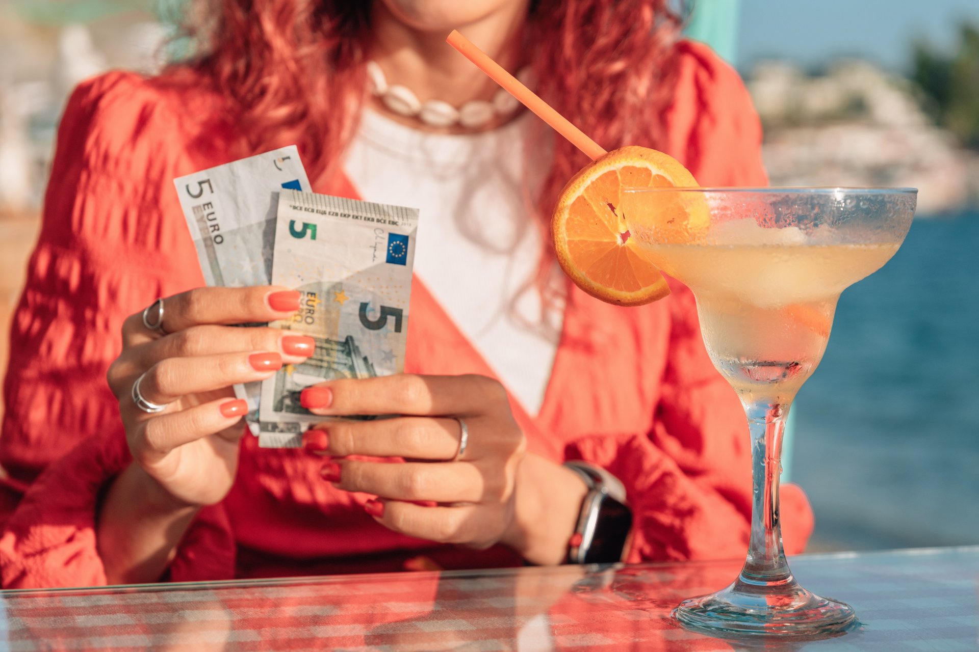 Femeie aflata la o cafenea de pe plaja care numara bancnote euro, in timp ce pe masa are un cocktail, sugerand planificarea bugetului pentru o vacanta relaxanta la mare.