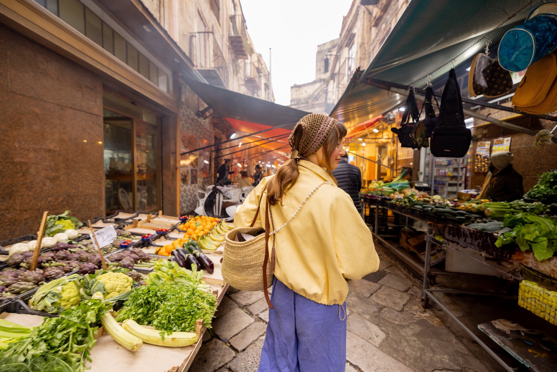 O femeie imbracata cu o geaca galbena alege salata proaspata dintr-o piata in aer liber din Sicilia, tinand telefonul in mana, inconjurata de tarabe cu legume si produse locale.