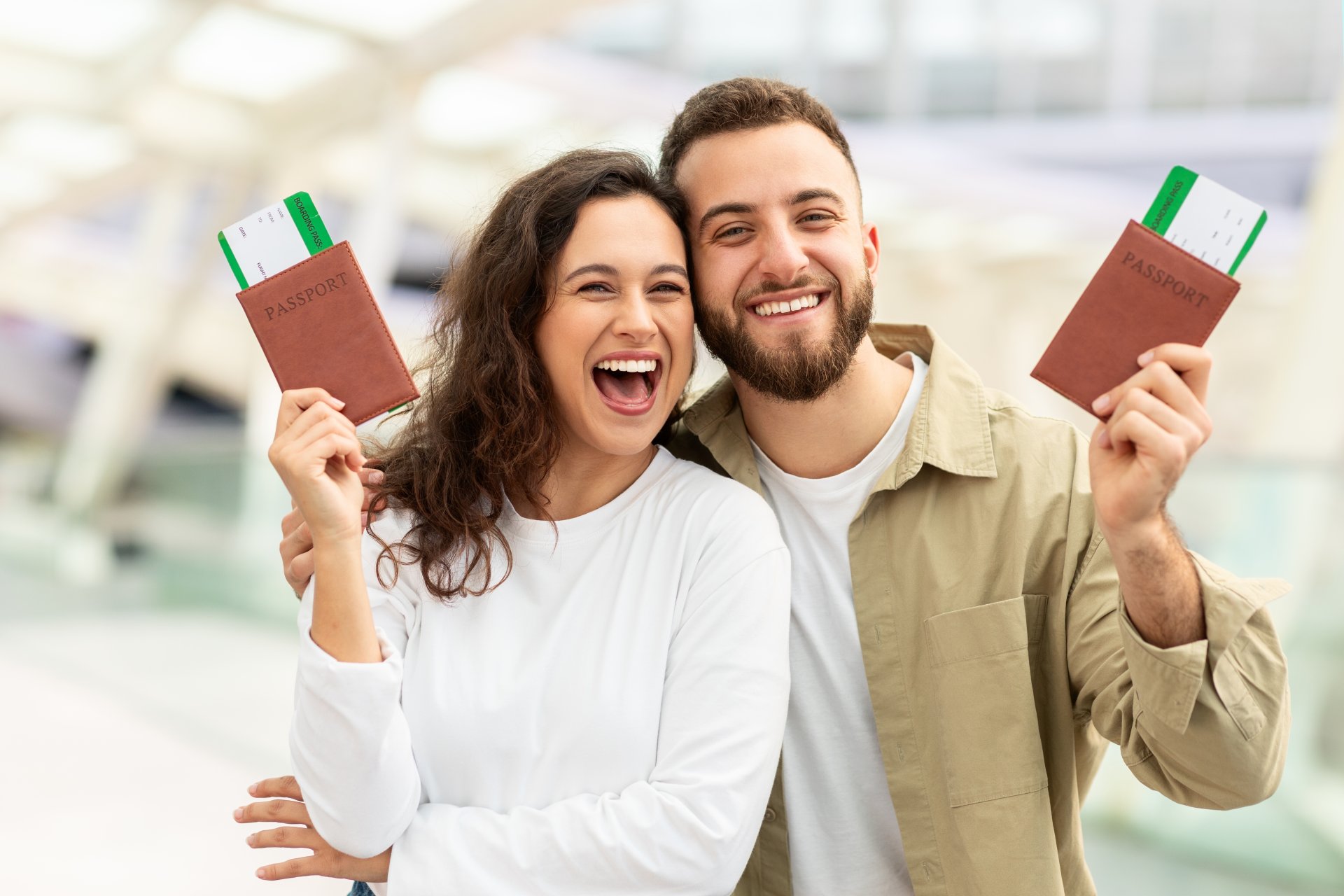 Un cuplu zambitor tine pasapoartele in mana intr-un aeroport si priveste spre camera, pregatit pentru plecare